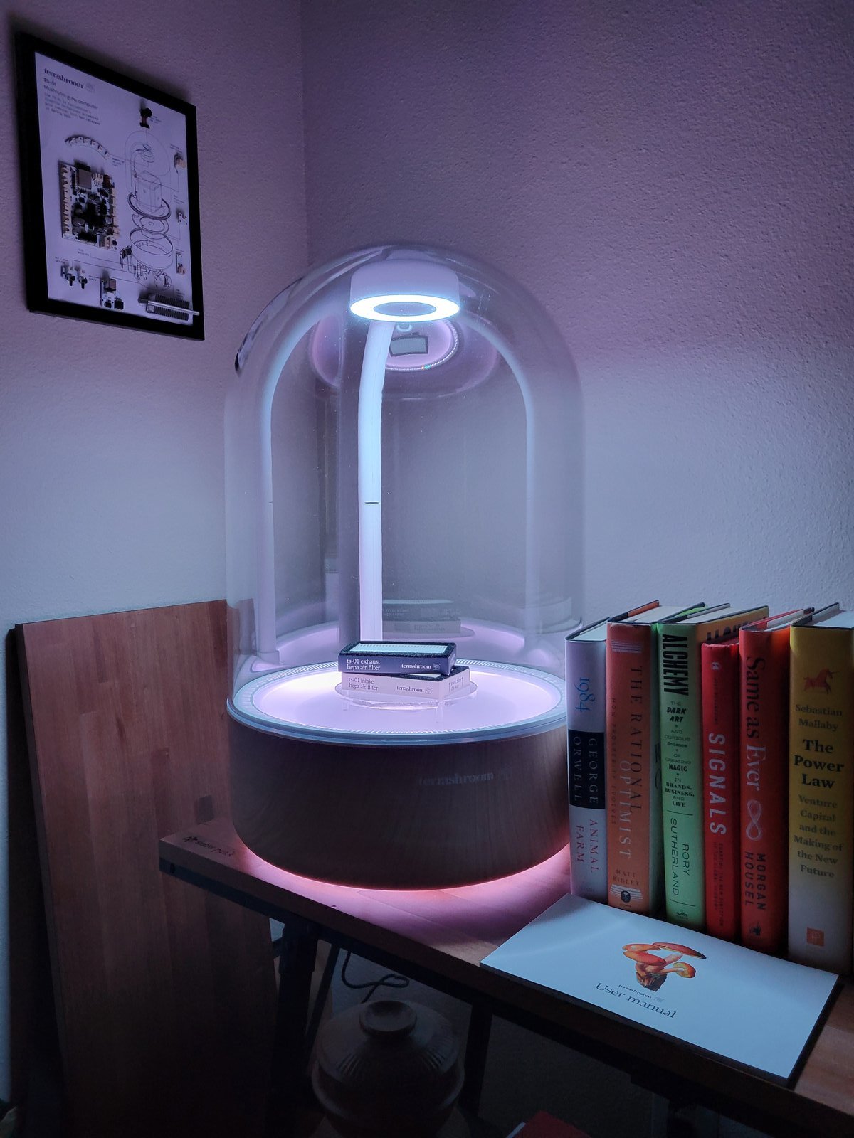 A terrashroom TS-01 grow computer on a shelf beside books and a framed technical drawing of the device